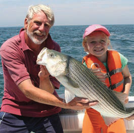 Man and small smiling boy hold very large striped bass on charter fishing boat